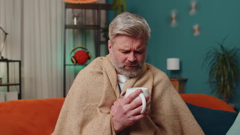 Sick-unhealthy-man-wrapped-in-plaid-sit-alone-shivering-from-cold-on-sofa-drinking-hot-tea-at-home
