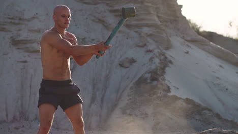 Bald-man-strongman-hits-a-hammer-on-a-huge-wheel-in-the-sandy-mountains-in-slow-motion.-strength-and-endurance-training-for-wrestlers.