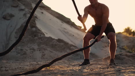 Athletic-man-on-exercise-around-the-sand-hills-at-sunset-hits-the-rope-on-the-ground-and-raised-the-dust.