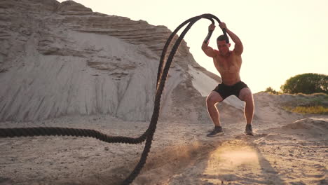Male-athlete-push-UPS-on-the-beach-and-hits-the-ground-with-a-rope,-circular-training-in-the-sun-on-a-sandy-beach.
