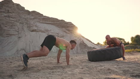Two-muscular-open-chested-athletes-train-in-active-mode-on-the-beach-doing-push-UPS-and-pushing-a-huge-wheel-against-a-sandy-mountain-at-sunset