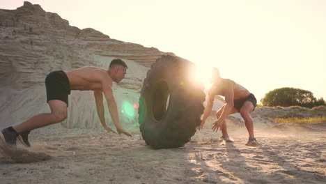Zwei-Muskulöse-Sportler-Mit-Offener-Brust-Trainieren-Im-Aktiven-Modus-Am-Strand,-Machen-Liegestütze-Und-Schieben-Bei-Sonnenuntergang-Ein-Riesiges-Rad-Gegen-Einen-Sandigen-Berg