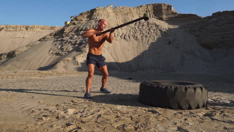 Muscle-athlete-strongman-man-hits-a-hammer-on-a-huge-wheel-in-the-sandy-mountains-in-slow-motion-at-sunset.-The-dust-from-the-wheels-rises.