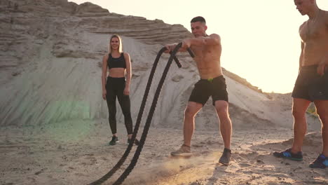 Group-crossfit-training-a-man-with-an-open-torso-doing-exercises-with-a-rope-a-woman-and-a-man-watching-the-performance-on-a-sandy-site.