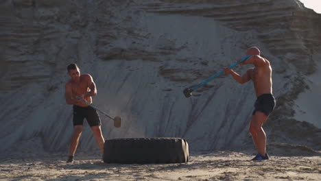Two-powerful-male-athletes-training-together-hit-the-wheel-with-a-hammer-at-sunset-in-the-desert.-Endurance-training