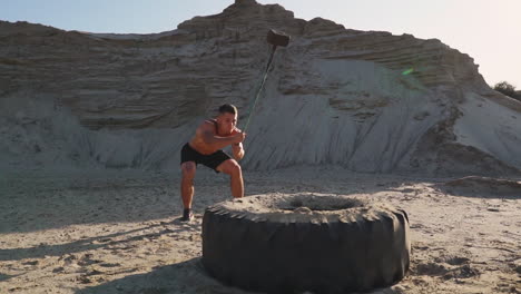 Muscle-athlete-strongman-man-hits-a-hammer-on-a-huge-wheel-in-the-sandy-mountains-in-slow-motion-at-sunset.-The-dust-from-the-wheels-rises.