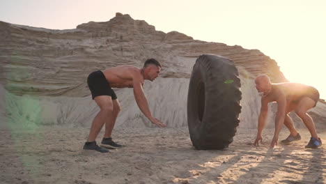 Two-athletes-train-in-active-mode-on-the-beach-doing-push-UPS-and-pushing-a-huge-wheel