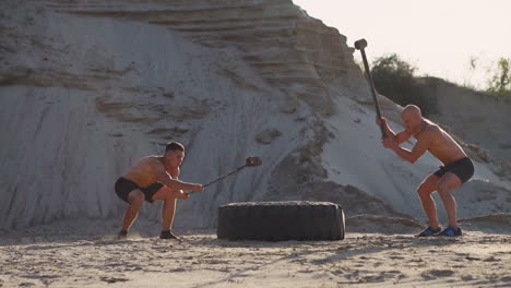 Two-male-athletes-training-together-hit-the-wheel-with-a-hammer-at-sunset-in-the-mountains-on-the-sand.-Endurance-training