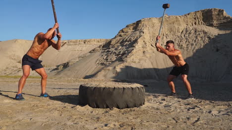 Two-powerful-male-athletes-training-together-hit-the-wheel-with-a-hammer-at-sunset-in-the-desert.-Endurance-training