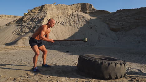 Bald-man-strongman-hits-a-hammer-on-a-huge-wheel-in-the-sandy-mountains-in-slow-motion.-strength-and-endurance-training-for-wrestlers.