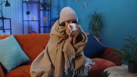 Sick-unhealthy-woman-wear-hat-wrapped-in-plaid-sit,-shivering-from-cold-on-sofa-drinking-hot-tea