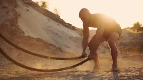 Male-athlete-doing-push-UPS-on-the-beach-and-hitting-the-rope-on-the-ground,-circular-training-in-the-sun-on-a-sandy-beach-raising-dust-in-slow-motion.