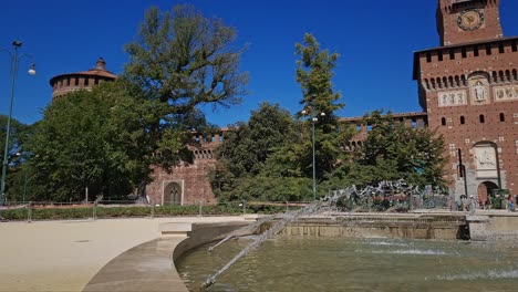 Agua-Pulverizada-Desde-La-Fuente-De-Piazza-Castello-Mientras-Los-Visitantes-Entran-Y-Salen-Del-Castillo-Sforza