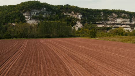 Aerial-dolly,-fresh-sown-soil-field-at-base-of-rocky-valley,-clean-patterned-tractor-lines