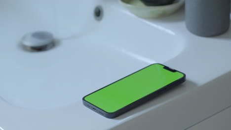 Kreativer-Einsatz:-IPhone-14-Mit-Grünem-Bildschirm-Auf-Dem-Waschbecken-Im-Badezimmer,-Tropfendes-Wasser-Aus-Dem-Wasserhahn,-Umgebungsbeleuchtung,-Chroma-Key
