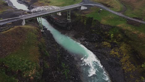 Drohnenaufnahme-Eines-Tobenden-Isländischen-Flusses-Und-Einer-Brücke-über-Den-Schwarzen-Felsen-Und-Stromschnellen