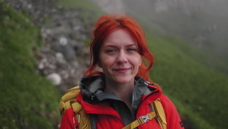 Slow-motion-right-panning-shot-of-a-smiling-hiker-girl,-she-has-red-hair-and-wears-an-orange-red-puffy-jacket-and-a-yellow-backpack