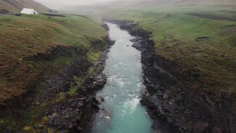 Drone-shot-of-a-raging-Icelandic-river-and-green-farm-fields-above-the-black-rocks-and-rapids