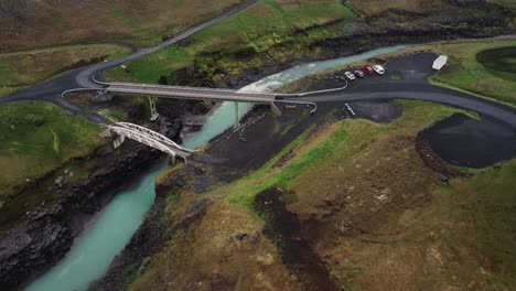 Drohnenaufnahme-Eines-Tobenden-Isländischen-Flusses-Und-Einer-Brücke-Mit-Geparkten-Autos-über-Den-Schwarzen-Felsen-Und-Stromschnellen