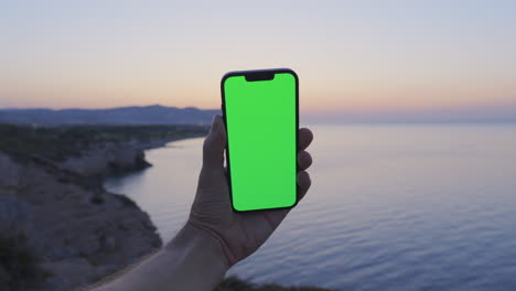 Golden-Hour-POV:-Man's-Hand-Holding-iPhone-14-with-Green-screen-On-Cliffside-near-Ocean,-Chroma-Key