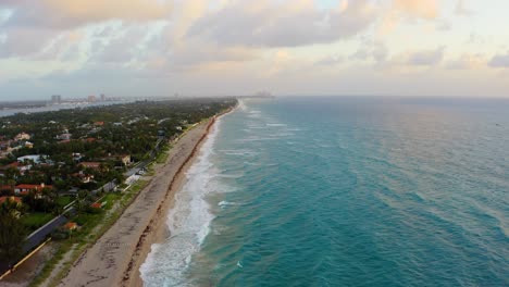 Aerial-drone-shot-slowly-flying-over-the-beach-on-Palm-Beach-while-the-waves-crash-and-the-sunrise-lights-up-the-clouds-and-island