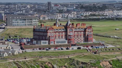 Static-aerial-shot-of-the-Headlands-Hotel-with-tourists-walking-past-towards-Fistral-Beach