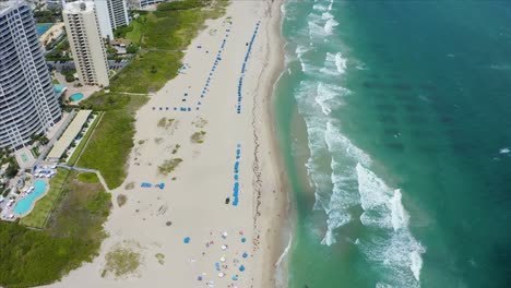 Aerial-drone-shot-flying-forward-over-umbrellas-and-tourists-on-Singer-Island-beach-near-the-Ritz-Carlton-residential-building-with-the-atlantic-ocean-waves-crashing-on-the-beach