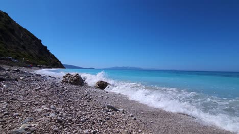 Walking-on-pebbles-of-paradise-beach-washed-by-white-sea-waves-on-beautiful-seaside-with-mountains-and-blue-turquoise-seawater-in-Albania