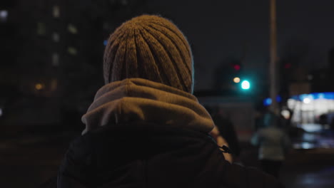 Rear-view-tracking-follows-man-in-layered-hoodie-sweatshirt-and-beanie-walking-through-downtown-city-scape-at-night