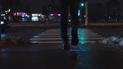 Low-angle-rear-view-follow-of-male-in-jeans-and-black-boots-crossing-city-street-at-night