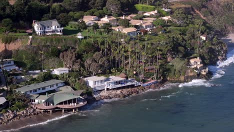 Luxury-beach-front-multi-million-dollar-homes-over-Malibu-Pacific-Ocean-coastline,-aerial-rising-on-picturesque-day-while-waves-crash-along-the-shore