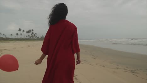 A-beautiful-woman-in-a-red-dress-holding-a-red-balloon-on-the-beach-looking-out-towards-the-ocean,-an-Indian-woman-with-balloons