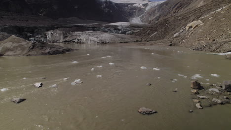 Drone-shot-of-dead-Pasterze-Glacier-lake-water-with-floating-iceberg,-Stream-of-muddy-glacier-water