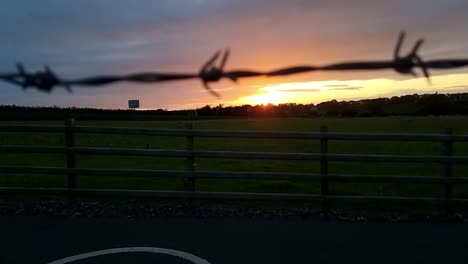 Looking-through-barb-wire-fence-to-sunset-orange-skyline-over-agricultural-farmland-field