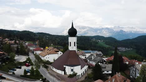 Rising-aerial-view-of-the-local-church-overlooking-Oberbozen,-Italy's-countryside