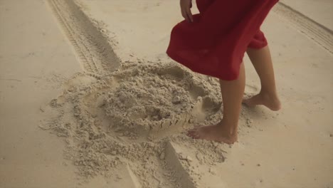 Eine-Frau-In-Einem-Roten-Kleid-Steht-Im-Sand,-Bückt-Sich-Dann,-Nimmt-Den-Sand-In-Ihre-Hände-Und-Spielt-Damit