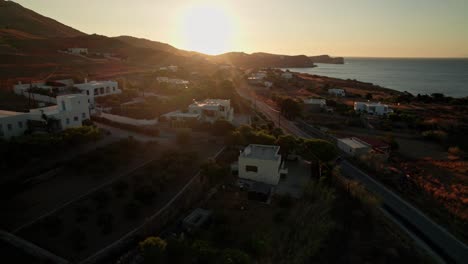 Quick-drone-whip-pan-towards-sunset-above-villas-on-greek-island-syros,-follow-motorbike-on-coastal-road