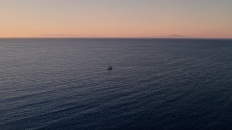 Drone-descends-to-boat-fishing-in-open-ocean,-no-one-else-in-sight,-golden-hour-sunset-sky,-ocean-ripples-at-perpendicular-angle