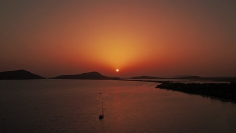Aerial-reverse-dolly-above-anchored-sailboat-as-ball-of-sun-sets-casting-warm-gradient-red-rays-into-sky-above-pylos-greece
