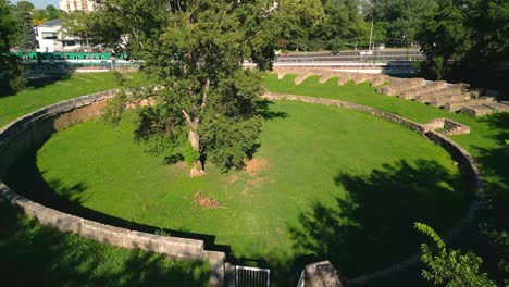 Tree-In-The-Middle-Of-Aquincum-Civil-Amphitheatre-With-Train-And-Traffic-In-The-Background-In-Budapest,-Hungary