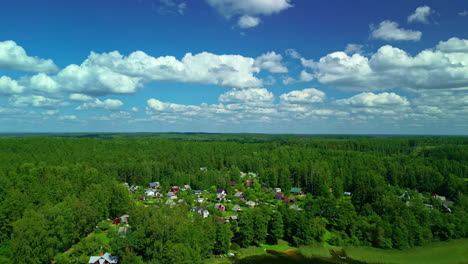 Cinematic-drone-shot-flying-over-a-forest-with-houses-in-between-them-on-a-clear-day-with-blue-sky-and-clouds