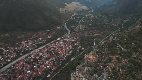 Drone-descends-above-greek-flagpole-and-cliff-to-reveal-town-of-leonidio-at-base-of-valley