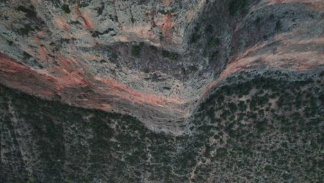 Drone-top-down-descends-across-intimidating-sheer-cliff-face-with-dry-shrub-landscape,-tilt-up-pullback-to-leonidio-greece