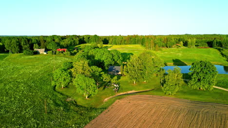 Toma-Aérea-Orbital-De-Una-Residencia-Rural-Entre-Campos-Verdes-Y-árboles-Bajo-El-Sol-De-Verano,-Letonia