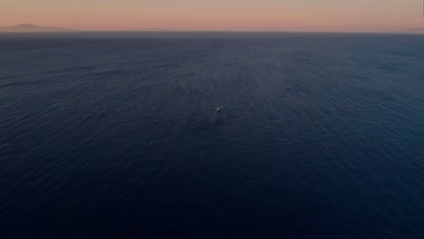 Single-boat-floats-alone-in-the-middle-of-large-blue-expanse-with-red-orange-gradient-sky-at-golden-hour
