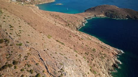 Aerial-bird's-eye-view-tilt-up-along-dry-coastline-of-syros-greece,-reveals-sailboat-and-peninsula