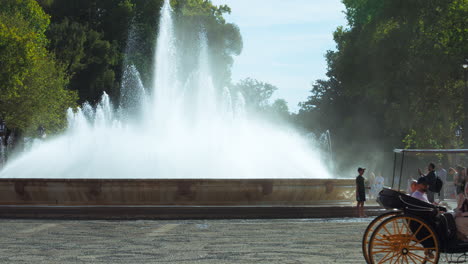 Fountain-at-Paza-España,-Seville,-with-tourists-nearby-and-horse-carriage-passing-by