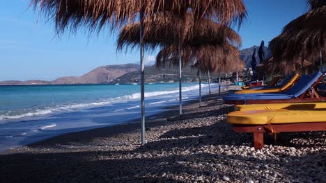 Playa-Tranquila-Con-Sombrillas-De-Paja-Y-Tumbonas-En-Albania,-Agua-De-Mar-Turquesa-Y-Olas-Blancas-Lavando-Guijarros,-Fondo-De-Vacaciones-De-Verano