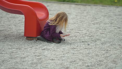 A-blonde-haired-girl-sits-near-the-slide-on-the-modern-outdoor-playground-and-plays-with-stones