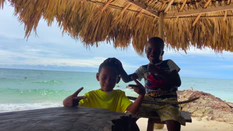 Two-black-kids-posing-and-smiling-to-camera-under-a-thatched-roof-on-a-tropical-beach